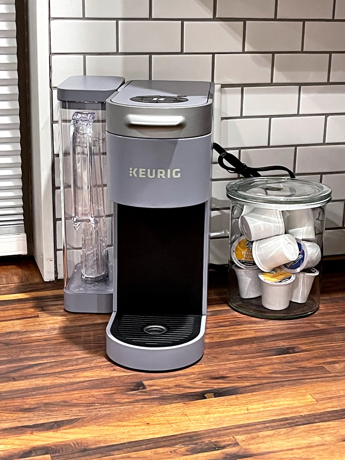 A modern coffee maker is positioned beside a glass jar filled with coffee pods. The machine features a sleek design with a prominent water reservoir, and a textured black panel is visible in the front. The background includes light-colored subway tile, enhancing the kitchen ambiance.