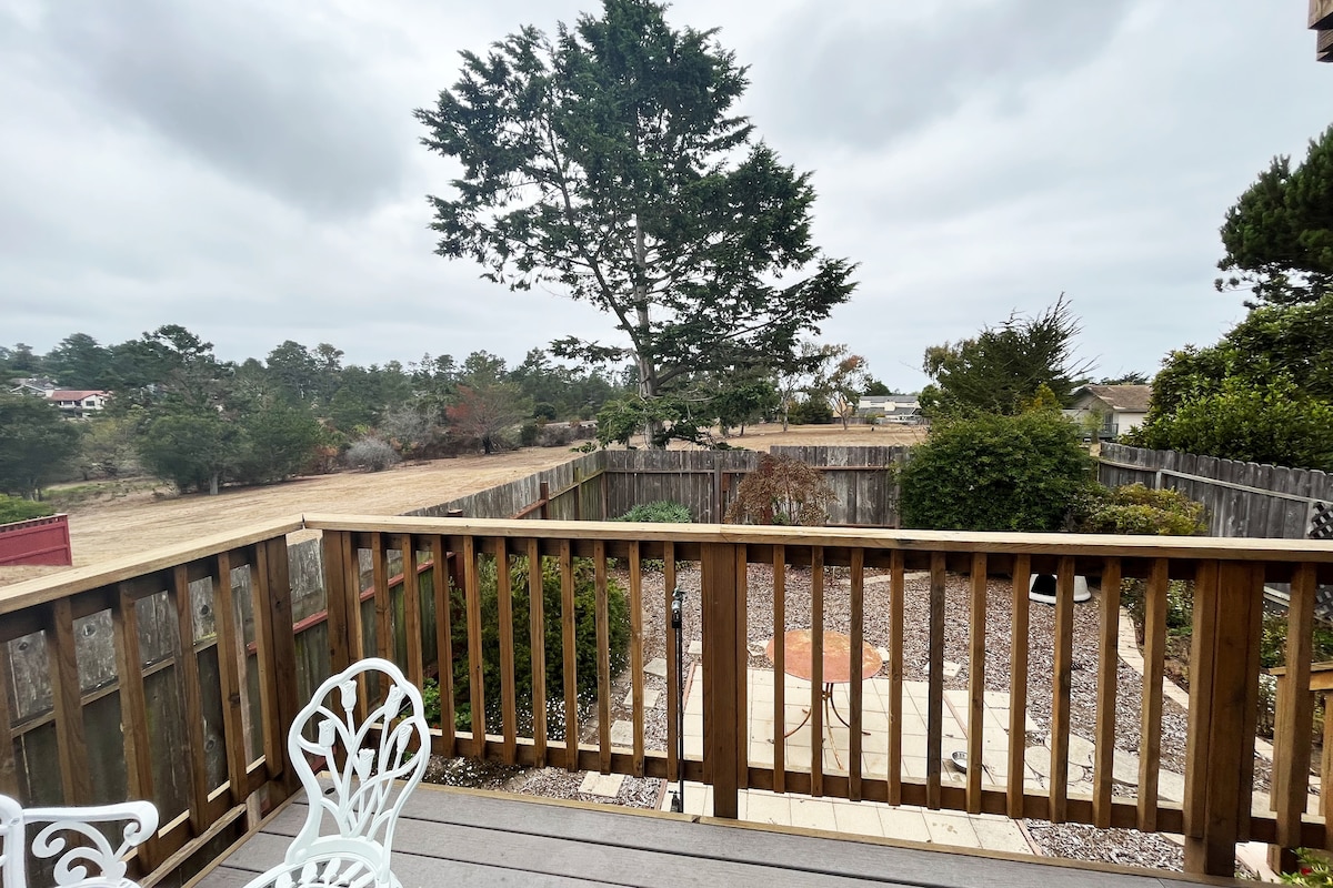 A spacious outdoor deck is featured, with white chairs arranged on one side. Beyond the railing, a serene garden area is visible, lined with a wooden fence and diverse greenery. A large tree stands prominently, surrounded by a tranquil landscape under an overcast sky.