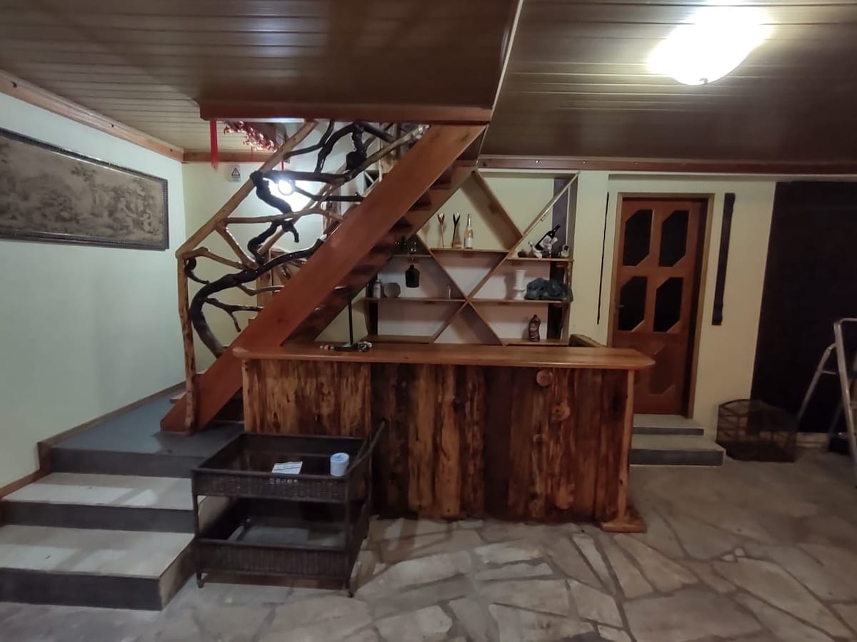 A rustic wooden bar counter is placed at the entrance, complemented by a unique staircase with a natural tree branch design. Stone tiles cover the floor, and simple wooden shelves display various items. A wooden door offers access to another area.