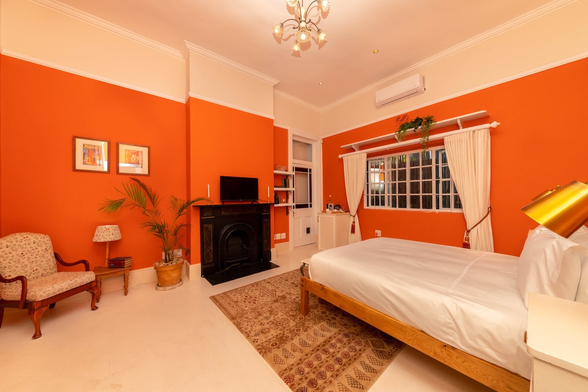 A spacious room features warm orange walls and high ceilings accentuated by a chandelier. A cozy seating area includes a patterned chair and side table. A modern TV rests atop a unit near a decorative fireplace, while soft light streams through large windows with curtains.