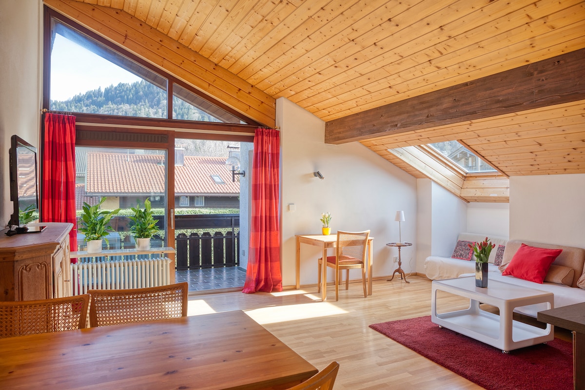 A bright and airy living space features a wooden ceiling with large windows allowing natural light to fill the room. A cozy sofa with decorative pillows is positioned near a coffee table, and a dining area with a wooden table and chairs is visible. Red curtains add a pop of color.