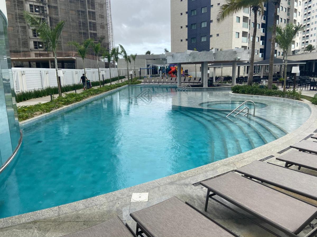 A modern pool area is highlighted with a gently curving shape, surrounded by lounge chairs. Palm trees line the edges, and nearby structures provide a contemporary backdrop. The water appears clear and inviting, reflecting the cloudy sky above.