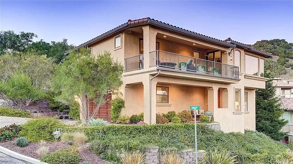 A two-story villa features a warm exterior with a tiled roof and expansive balcony. Lush landscaping surrounds the home, with various plants and shrubs enhancing the natural setting. The inviting entrance showcases a well-maintained pathway leading to the front door.