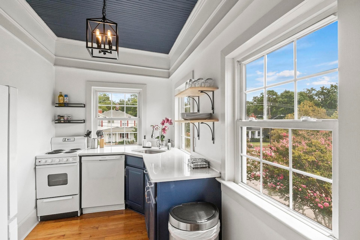 A well-equipped kitchen features white cabinetry and a dark blue island, complemented by a window overlooking vibrant outdoor greenery. The area is illuminated by a hanging light fixture, and essential appliances including a stove and dishwasher are neatly positioned for convenience.