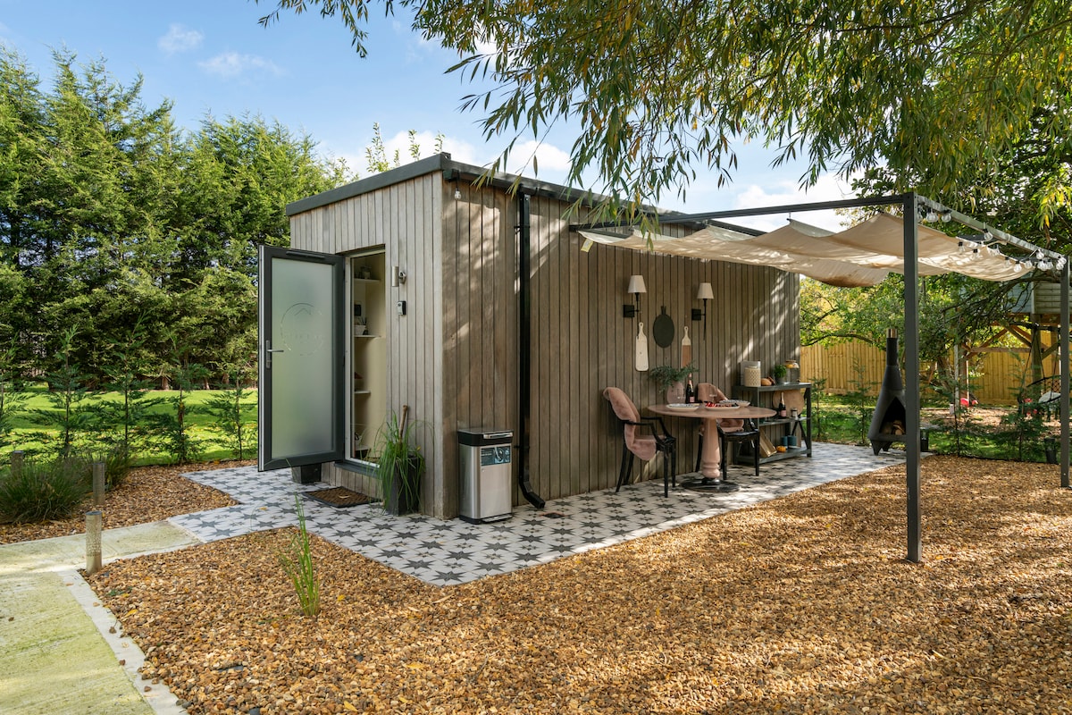The exterior of the cedar pod is showcased with a covered dining area featuring a table and chairs. A decorative stone path leads to the glass entrance, surrounded by landscaped greenery and woodchip bedding. Shade is provided by an overhead canopy.