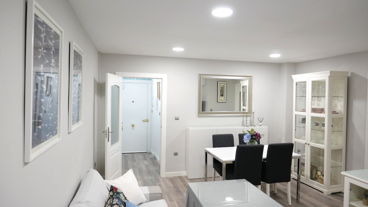 The living area features a light-colored sofa and a dining table surrounded by four chairs. Artwork in frames adorns the walls, and a glass-fronted cabinet displays decorative items. Soft overhead lighting brightens the space, creating a welcoming atmosphere.