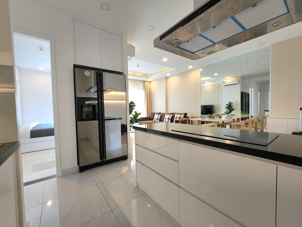 A modern kitchen is shown, featuring sleek white cabinetry and a dark countertop. Stainless steel appliances, including a refrigerator, are visible. The open layout connects to a dining area with a wooden table and chairs, enhancing the airy atmosphere of the space.
