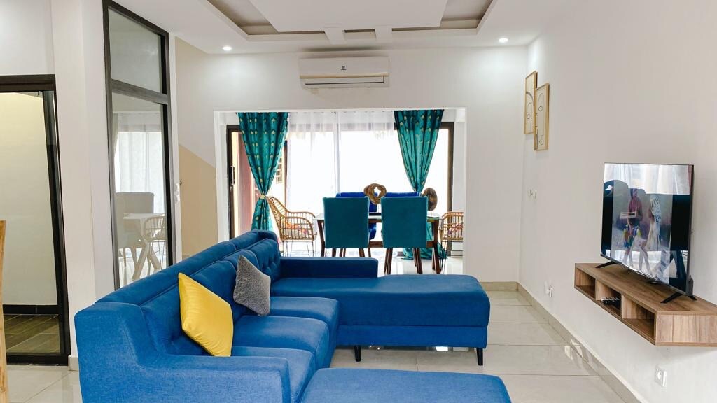 A modern living area features a large blue sectional sofa, complemented by decorative cushions. Natural light enters through expansive glass doors, leading to a dining space furnished with stylish chairs. A flat-screen TV is mounted on the wall opposite the seating arrangement.