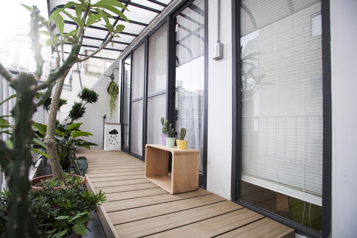 A wooden deck leads to large glass doors, providing access to the interior. Potted plants, including cacti, are placed on a modern wooden table, while additional greenery lines the walkway, creating a serene outdoor space. Natural light filters through the glass panels.