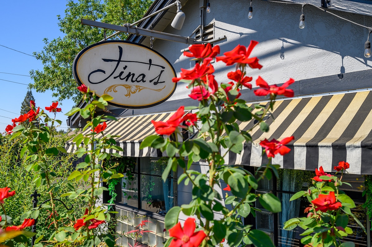 A charming exterior view of a building with a striped awning, showcasing a sign that reads 'Tina's.' Vibrant red roses are in full bloom, framing the entrance and adding a touch of color to the welcoming facade.