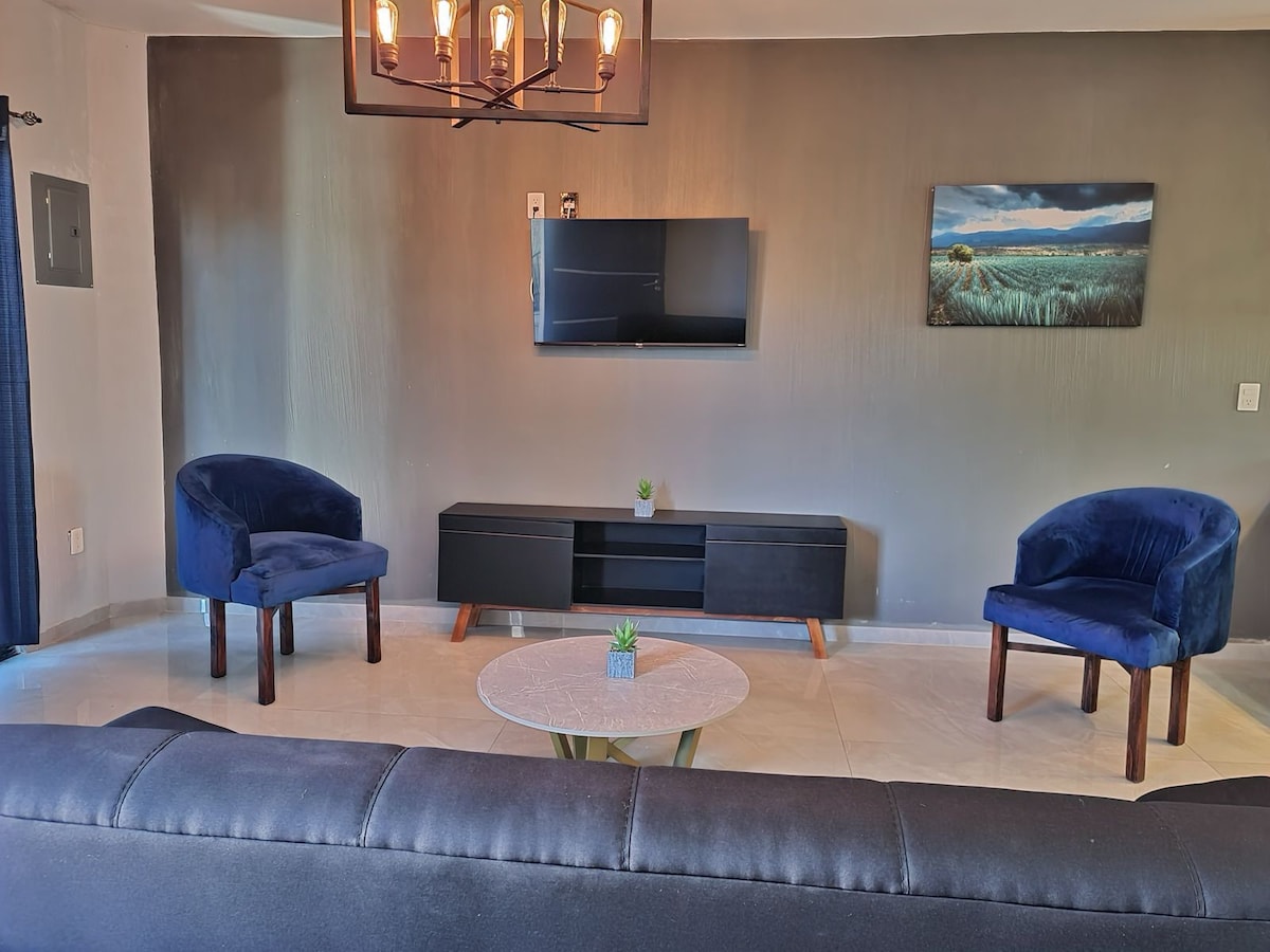 A modern living area features a dark sofa facing a mounted flat-screen television. Two plush blue chairs are positioned beside a round coffee table, with a small potted plant in the center. Soft lighting is provided by an overhead chandelier.