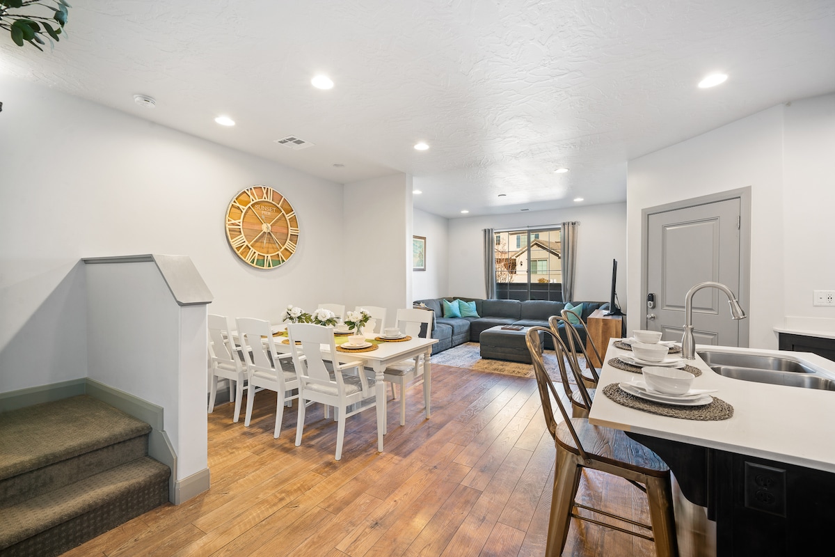 An open-concept living space features a dining table for six, complemented by white chairs. A spacious sectional sofa occupies one side, with natural light streaming in through large windows. The kitchen, equipped with modern appliances, maintains a clean and organized layout, while wood flooring creates a warm ambiance.