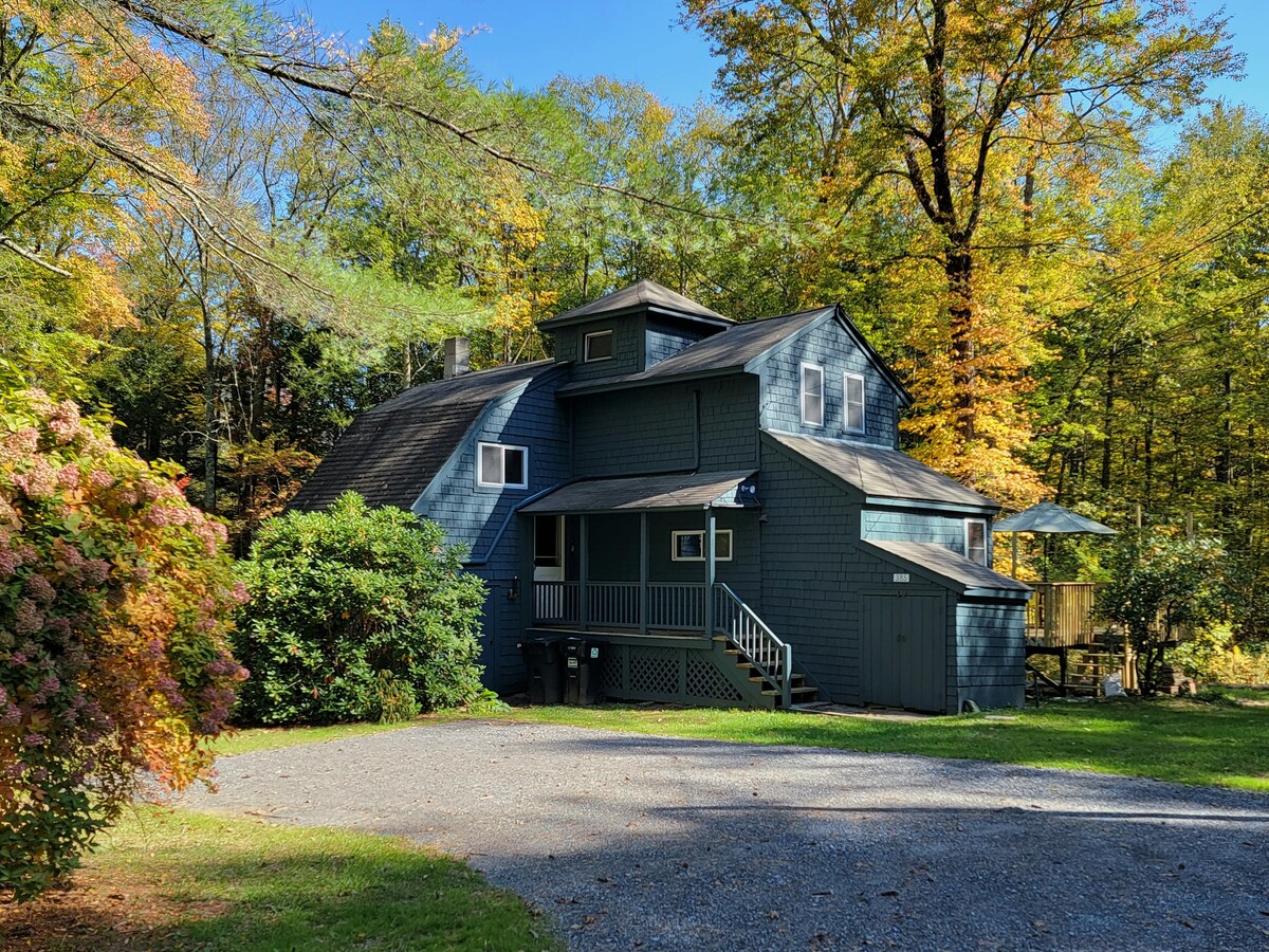 A charming blue cottage is nestled among vibrant autumn foliage. The two-story structure features a mix of gabled roofs and a covered porch. Lush greenery surrounds the gravel driveway, enhancing the serene setting.