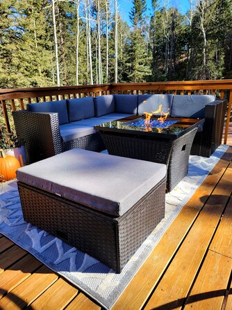 An outdoor seating area features a dark wicker sectional sofa with grey cushions and a faux fire pit table at its center. A cozy ottoman complements the seating, all arranged on a wooden deck surrounded by trees, creating a serene retreat.