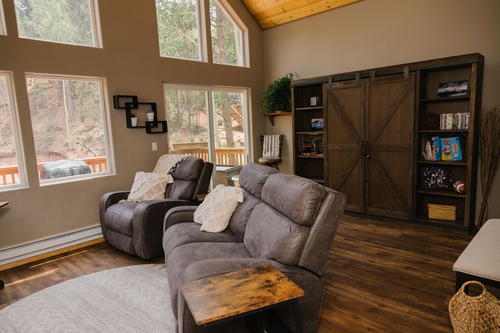 A comfortable living area features two reclining armchairs facing large windows that provide ample natural light. A wooden bookshelf adds a rustic touch, while a soft area rug anchors the space. The warm wood ceiling complements the serene mountain setting visible outside.
