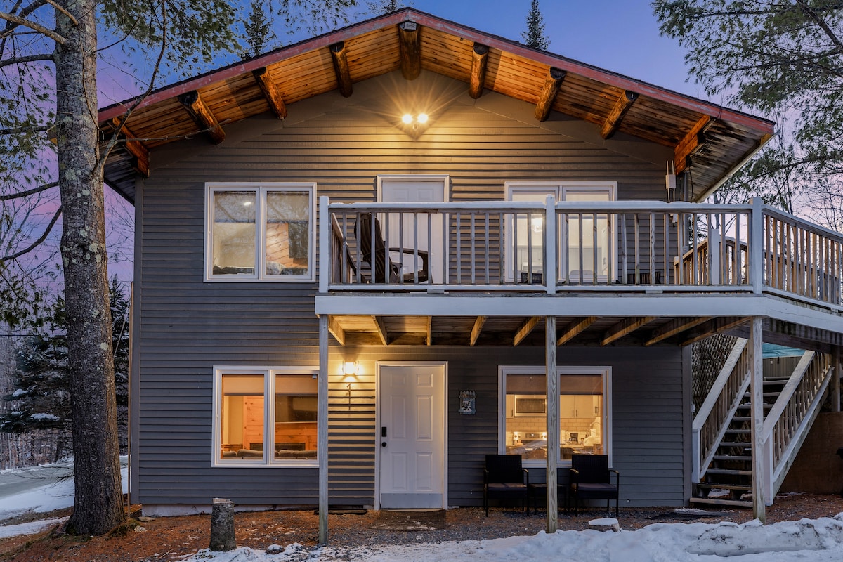 The two-story exterior of the chalet is shown with a welcoming porch featuring seating. Soft lighting highlights the entrance and upper deck, surrounded by trees. A snow-covered path leads to the front door, and large windows provide glimpses of the interior.