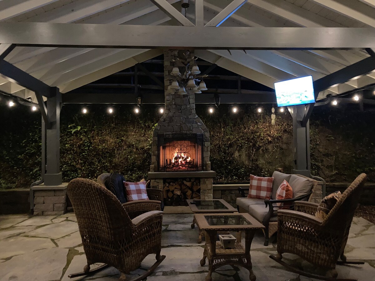 An outdoor pavilion features a stone fireplace, surrounded by cozy seating arrangements, including two armchairs and a coffee table. Soft lighting from hanging bulbs enhances the space, while a television mounted on the wall allows for entertainment during gatherings.