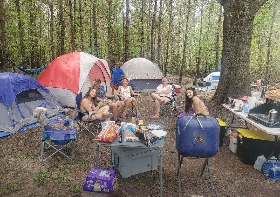 A group of individuals enjoys time around a campsite, surrounded by several colorful tents under tall trees. Portable chairs are set up, and a table displays snacks and a blue cooler, creating a casual gathering atmosphere in a wooded setting.