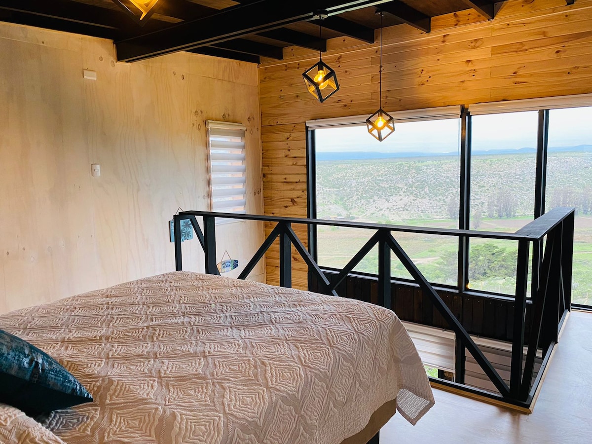 The loft features a spacious sleeping area with a light-toned quilted bedspread. Large windows provide expansive views of the surrounding landscape, while modern pendant lights hang above, illuminating the wooden accents of the room.