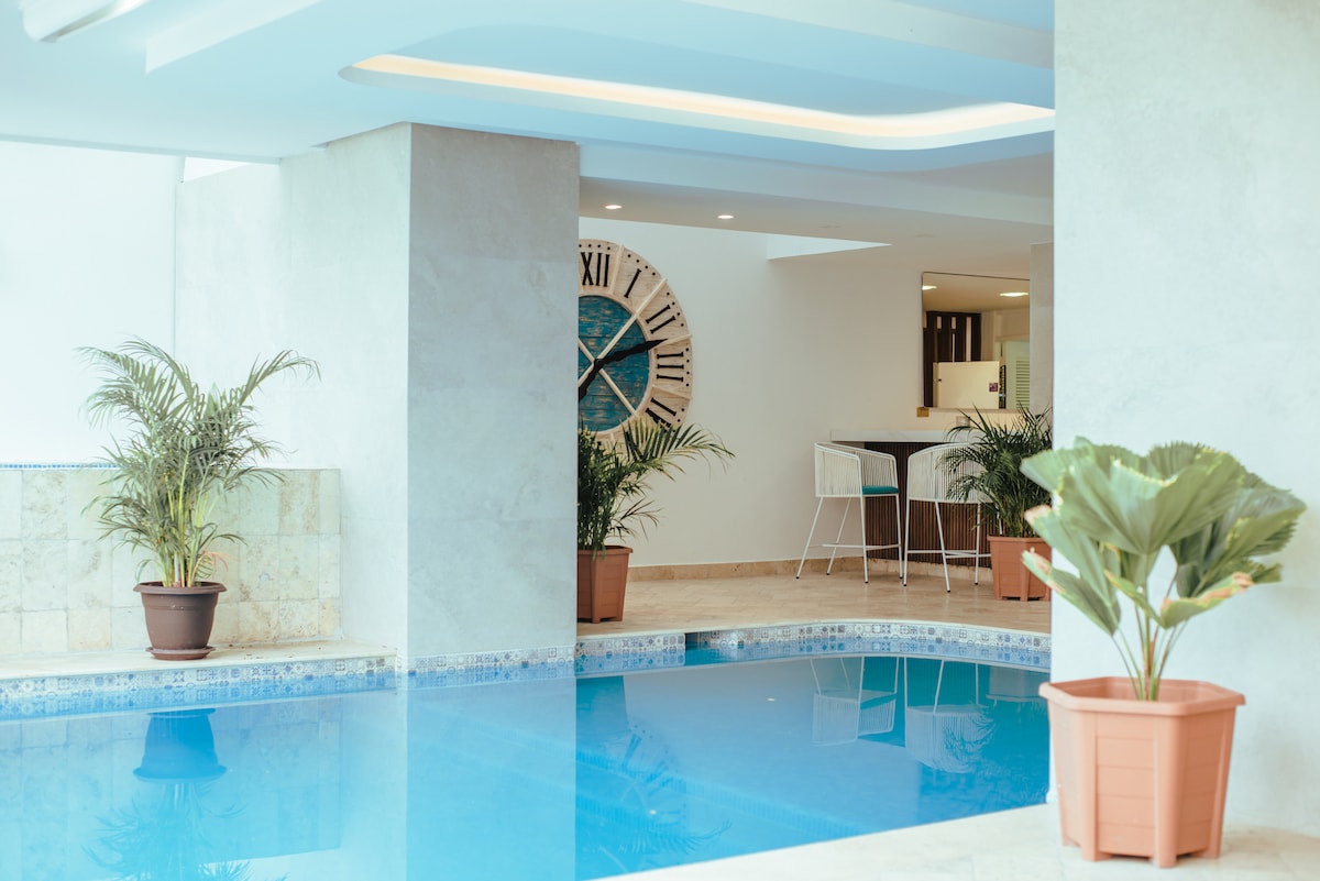 The indoor pool area features clear blue water surrounded by light-colored walls. Potted plants are placed around the pool, creating a serene environment. A large clock on the wall adds a modern touch, while a bar with high chairs is visible in the background.