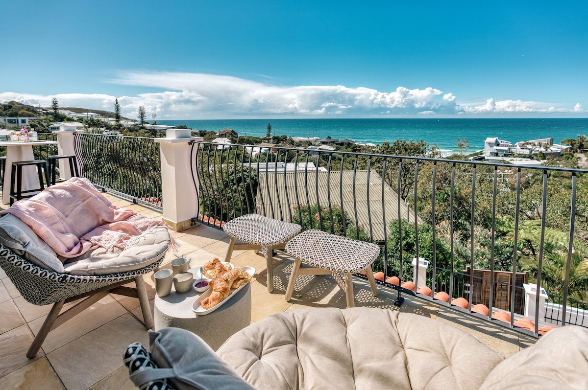An outdoor balcony is presented with airy seating, featuring plush cushions and a small table. Fresh pastries and two cups are arranged on the table, with expansive views of the ocean and distant clouds visible in the background.