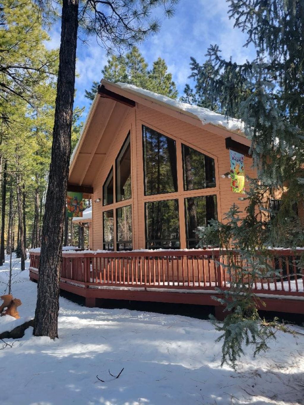 A spacious cabin features large glass windows that allow natural light to enhance the warm wooden exterior. A wide deck is visible, surrounded by snow-covered ground and tall pine trees, providing a peaceful wooded setting.