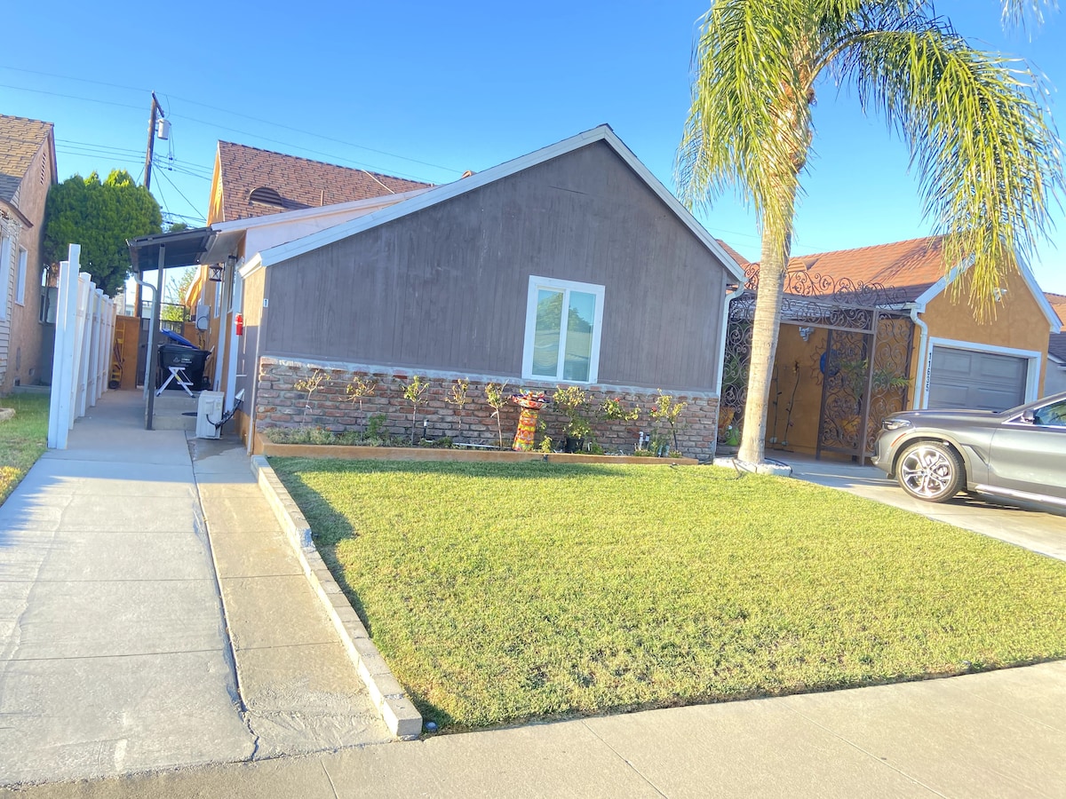 A single detached unit is shown, featuring a well-maintained exterior with a mix of stone and stucco. A neat lawn is visible in front, bordered by a concrete pathway and decorative plants. A palm tree adds a tropical touch beside the driveway.