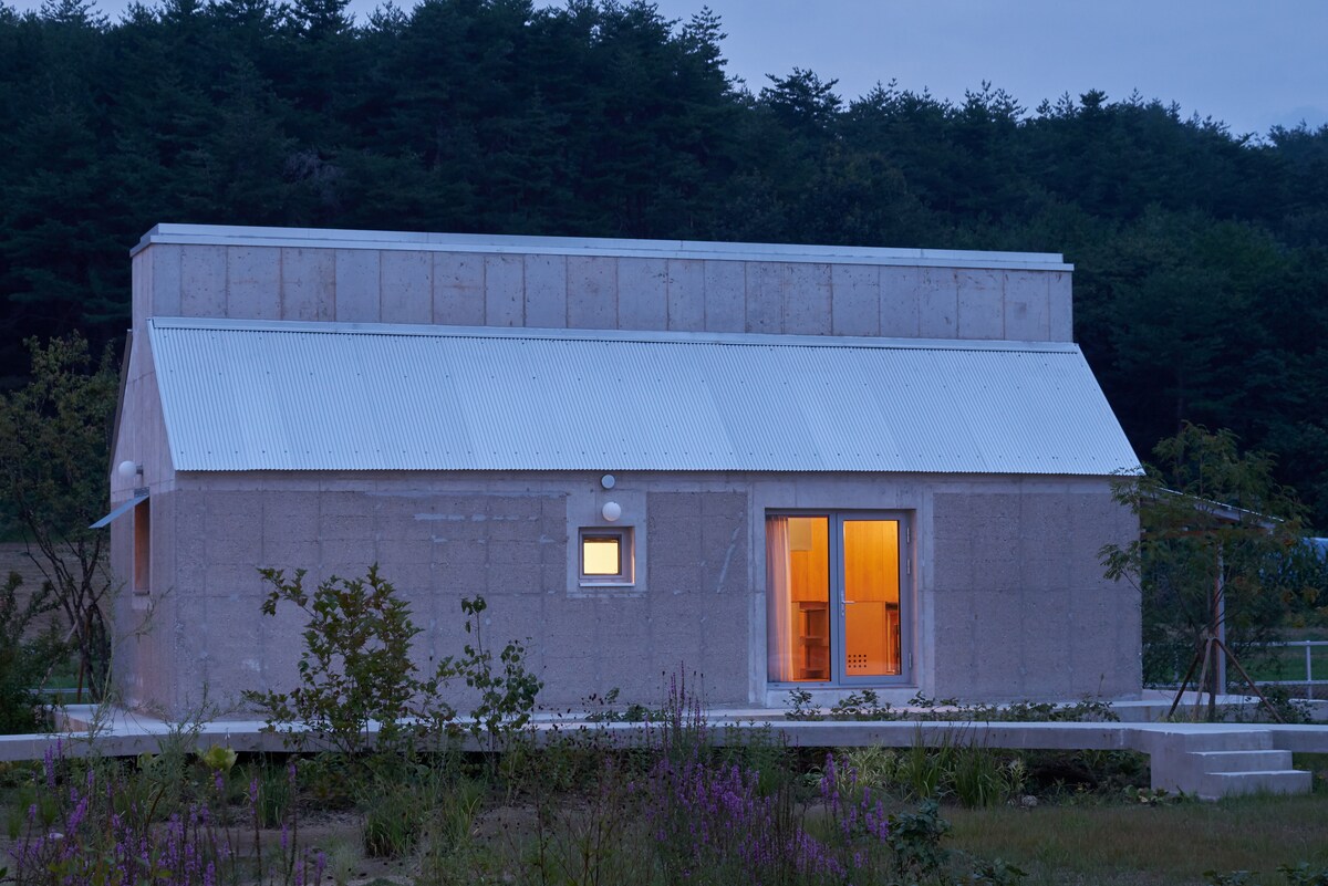 The exterior of a long, rectangular building is shown, featuring a metal roof and soft glowing lights from within. Surrounded by greenery, the building's façade is complemented by a small window and a pair of glass doors that open to the outdoors.