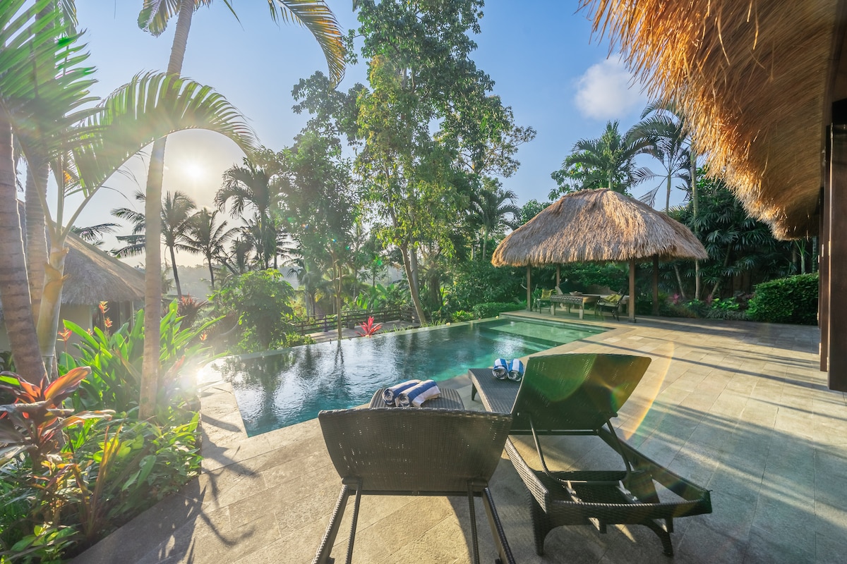 An infinity pool is framed by lush greenery and palm trees, reflecting the soft glow of the sun. Lounge chairs are positioned nearby, providing a serene spot for relaxation. A traditional thatched gazebo stands elegantly in the background, enhancing the tropical ambiance.