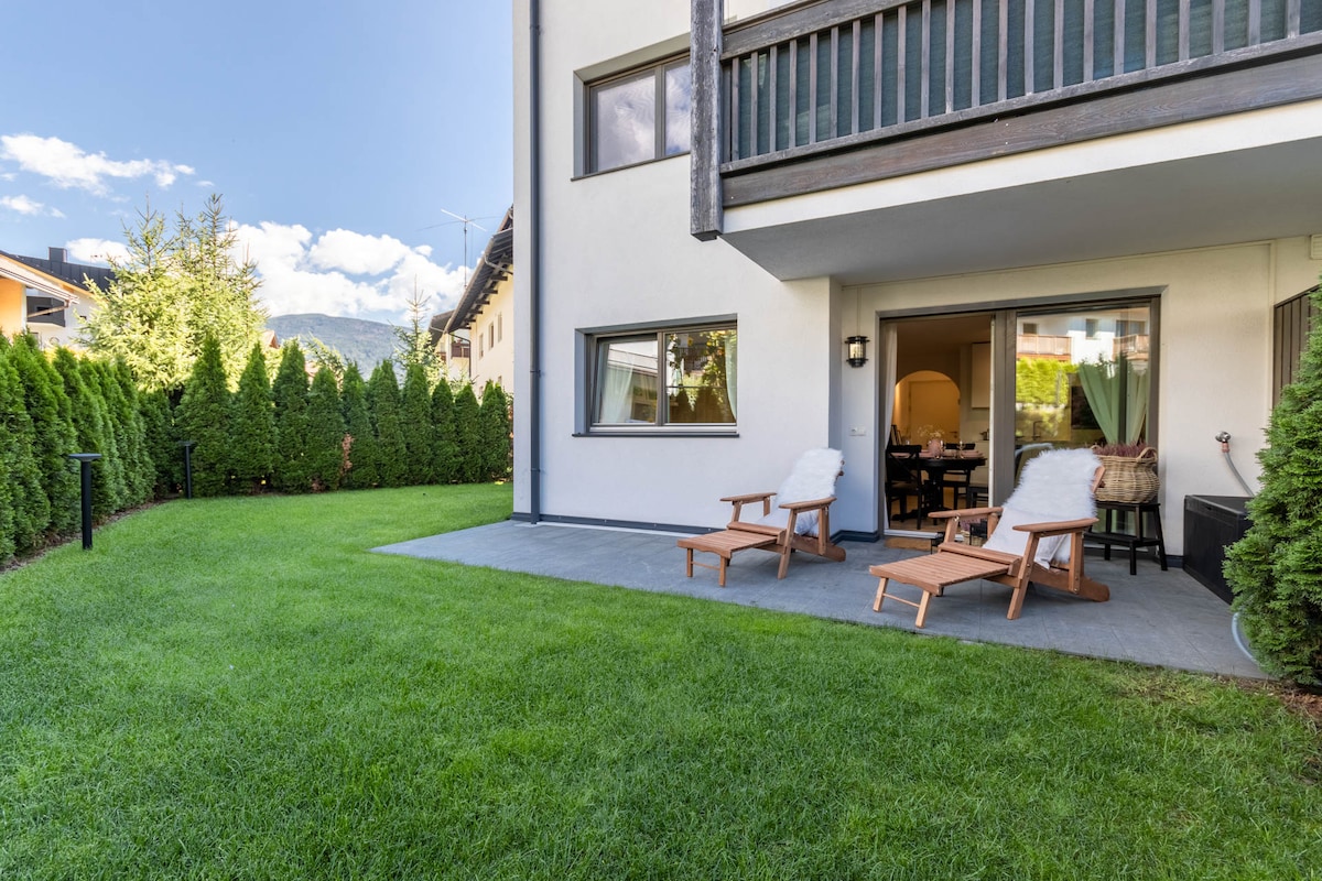 A serene garden space is presented with well-manicured grass, bordered by tall, lush hedges. Two wooden lounges are positioned on a paved area, inviting relaxation. The entrance to the interior is visible, framed by large glass doors that connect the outdoor area to the living space.