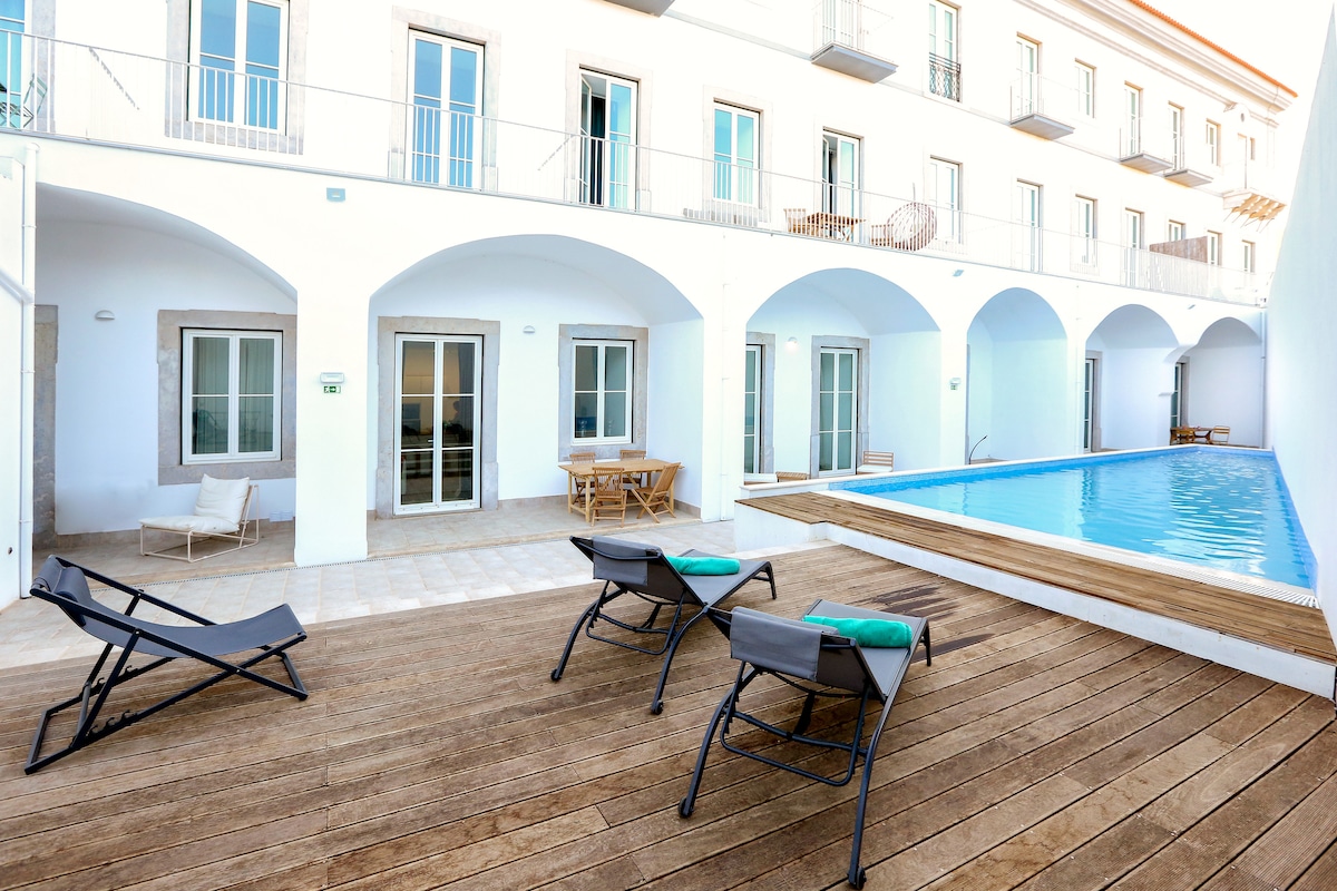 An outdoor area features a wooden deck with two lounge chairs positioned near a swimming pool. The pool is surrounded by a white wall and arches, with several windows visible in the background. A dining set is also present, adding to the inviting ambiance of the space.