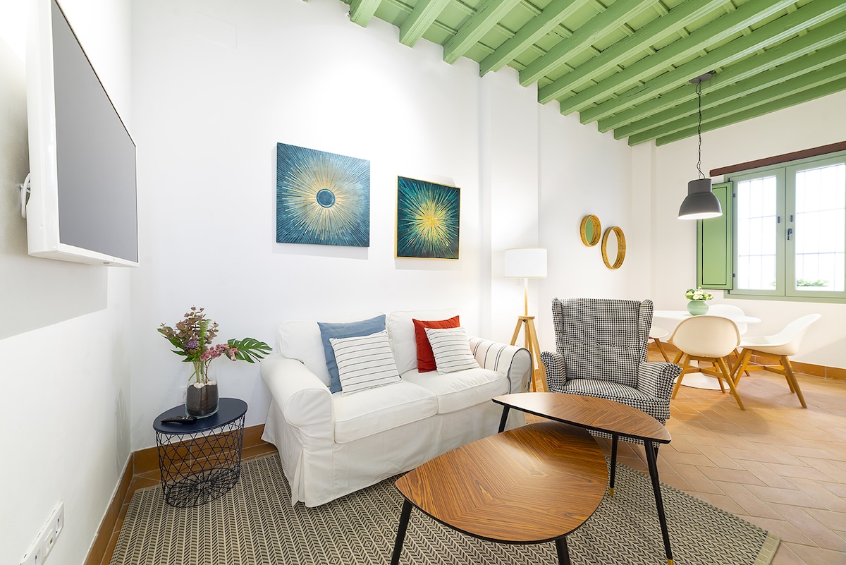 The living area features a white sofa accented with colorful cushions, accompanied by a textured armchair. Two circular tables are positioned in front, with artwork displayed on the walls. Natural light filters in through windows with green shutters, adding to the airy ambiance.