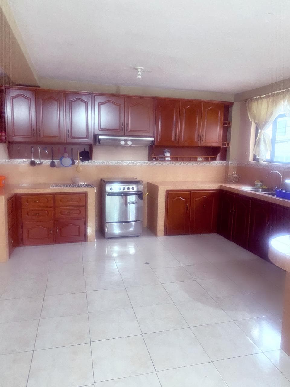 The kitchen features wooden cabinetry with a rich brown finish and ample counter space. A stainless steel stove and oven are positioned in the center, complemented by a large sink near a window. The tiled floor reflects light from the nearby window, creating a bright environment.