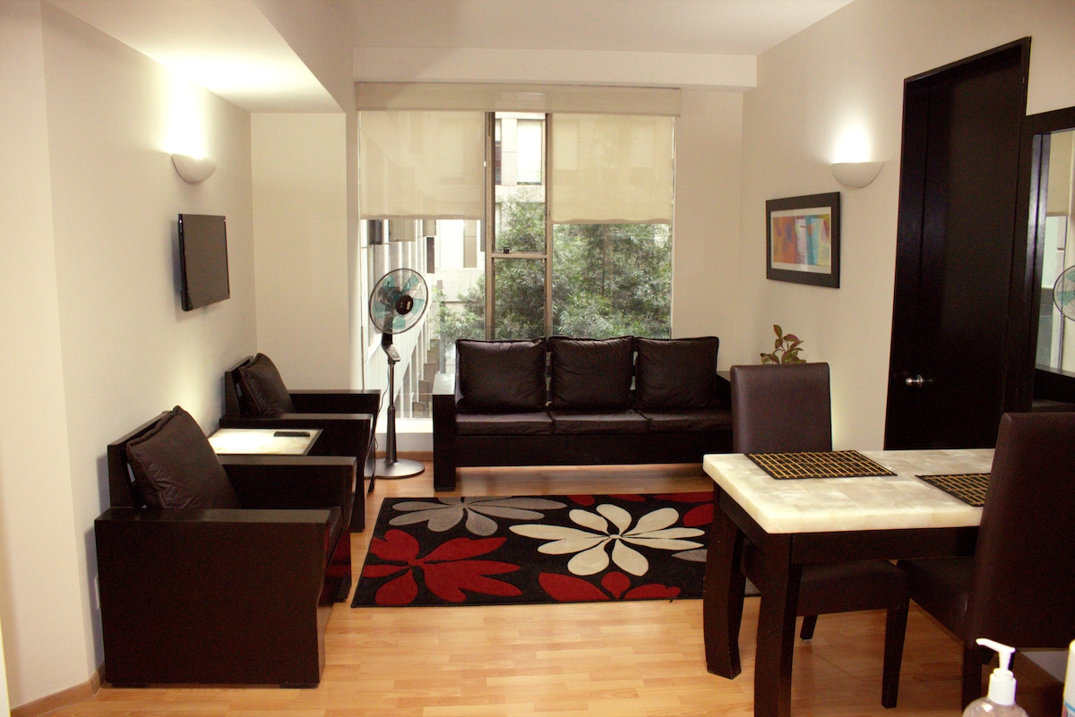 A modern living area features a stylish sofa and matching armchairs arranged around a decorative rug with floral patterns. A dining table with a stone top is positioned nearby, while large windows permit natural light into the space.