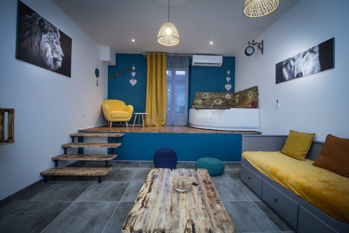 A spacious living area features a raised platform with a comfortable yellow couch and a wooden coffee table. Colorful poufs add a playful touch, while an inviting yellow armchair complements the blue accent wall. A jacuzzi is partially visible in the background beneath large windows.