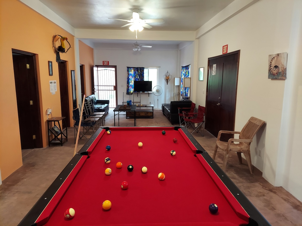 A vibrant leisure space is depicted, featuring a red tournament-sized pool table set for a game. Comfortable seating is arranged nearby, and a television is visible in the distance. Colorful curtains allow light to filter into the bright atmosphere.