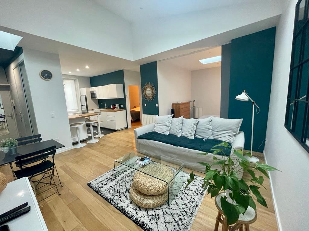 A spacious living area features a light-colored sofa centered on a textured black and white rug. A glass coffee table rests atop the rug, complemented by a potted plant nearby. The room is brightened by natural light from overhead skylights and a nearby window.