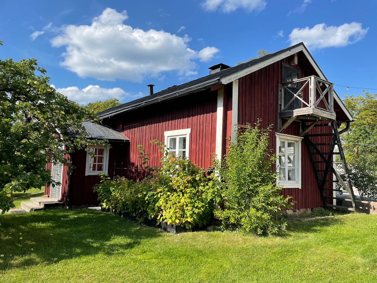 The exterior of a charming log cabin features rich red wood siding, surrounded by lush greenery and climbing plants. A staircase leads to an upper balcony, with white trim accents highlighting the windows. Blue skies and scattered clouds frame this inviting historical structure.