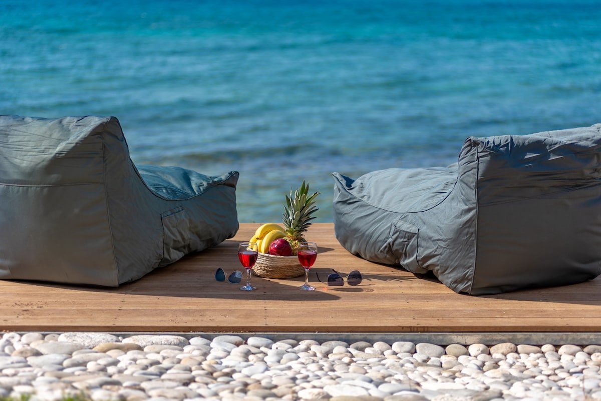 Two comfortable bean bags are positioned on a wooden deck facing the serene sea, complemented by a decorative bowl of fruits including bananas and a pineapple. Two red drinks in glasses sit on the deck, enhancing the relaxed beachfront setting.