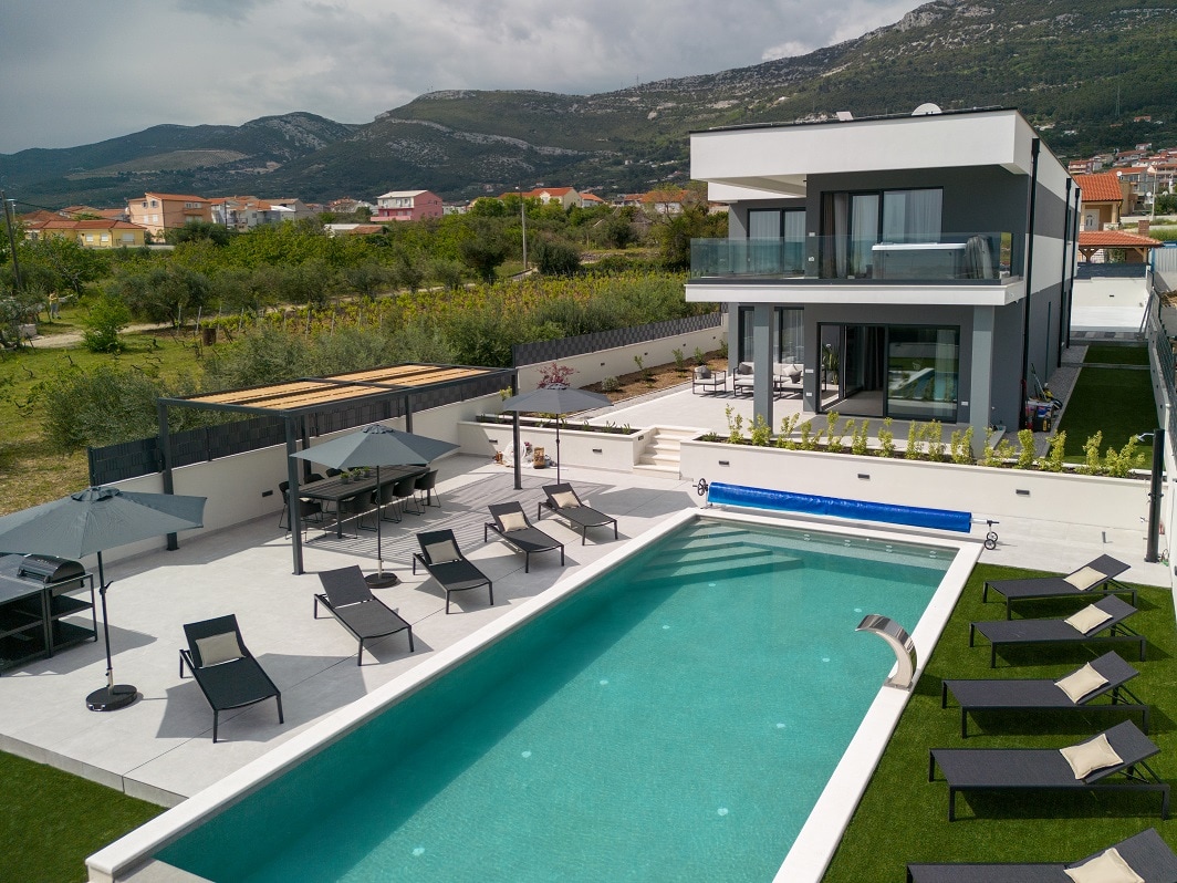 An aerial view of a modern villa features a large pool surrounded by lounge chairs. The outdoor area includes a dining space with a pergola and is bordered by lush greenery. The two-story villa displays multiple balconies overlooking the pool.