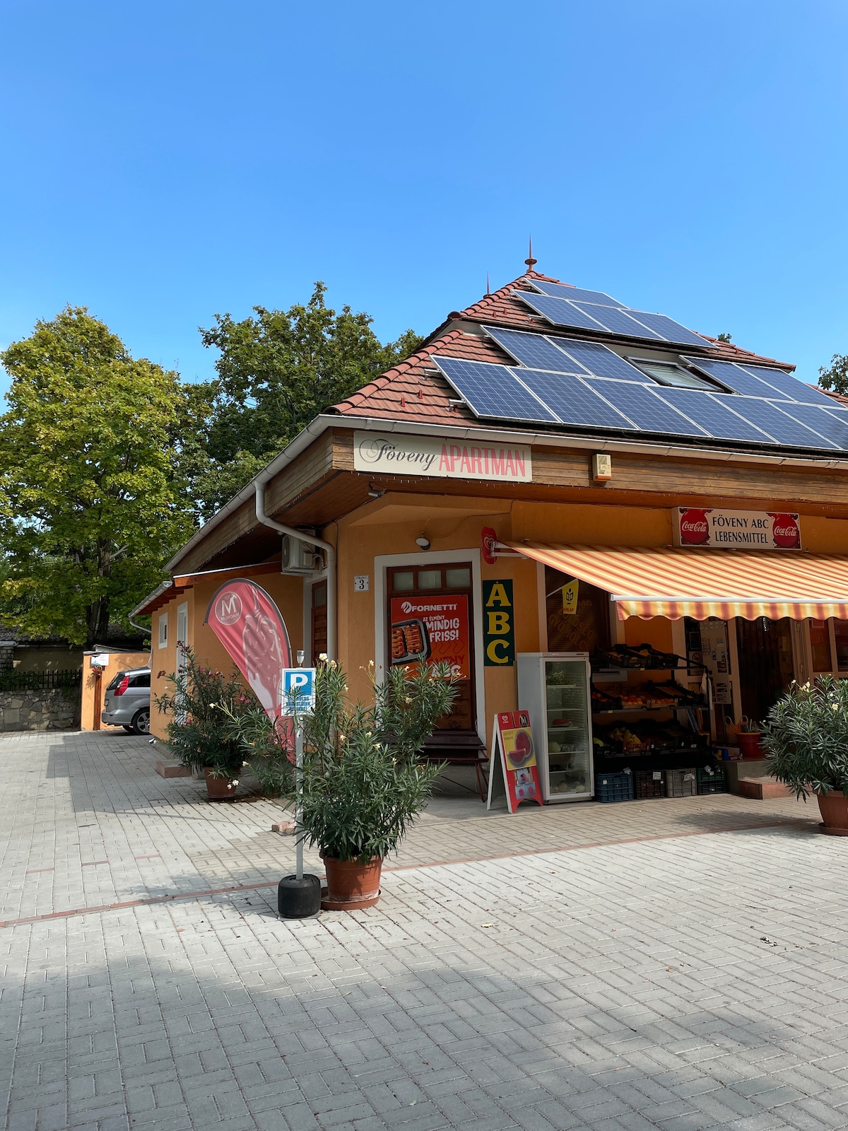 A quaint building with a sloped roof features solar panels for energy efficiency. The entrance displays a shop offering various goods, with bright signage and a striped awning. Potted plants flank the walkway, enhancing the inviting façade.