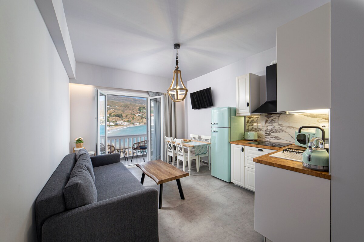An open-plan living area features a gray sofa and a wooden coffee table. A light-colored dining table and chairs are positioned nearby. Large sliding glass doors provide a view of the sea and lead to a balcony, enhancing the bright and airy space.
