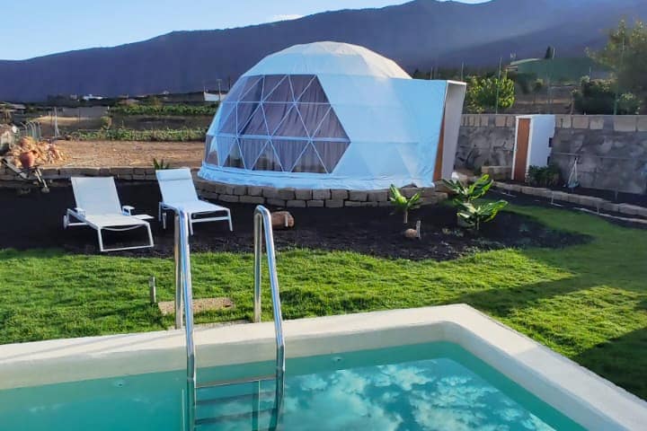 A modern dome structure is set against a backdrop of rolling hills and mountains. A private, heated swimming pool is visible in the foreground, with two lounge chairs positioned on the lawn, enhancing the outdoor space surrounded by greenery.