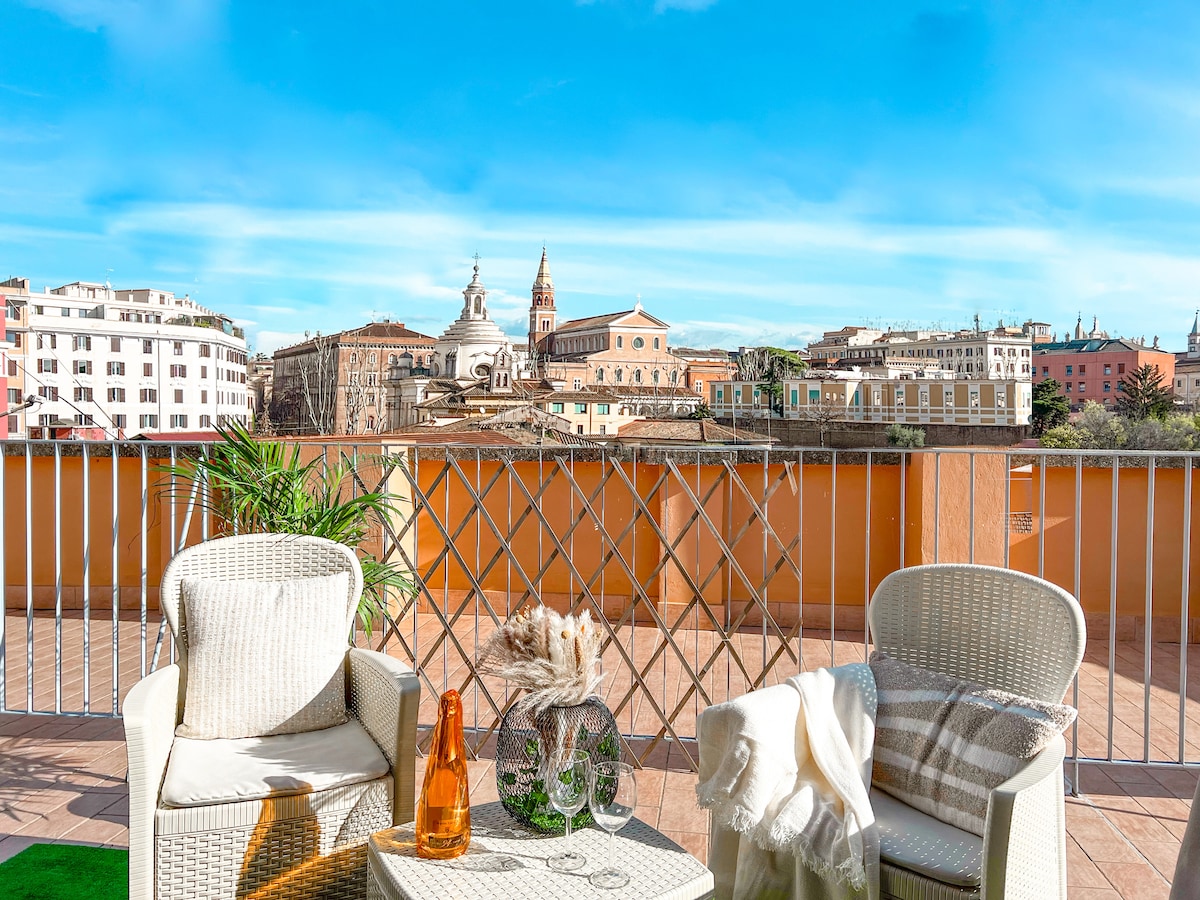 A cozy balcony features two light wicker chairs and a small table, accompanied by an amber bottle and decorative accents. A soft blanket is draped on one chair. The stunning view showcases historic buildings under a bright blue sky.