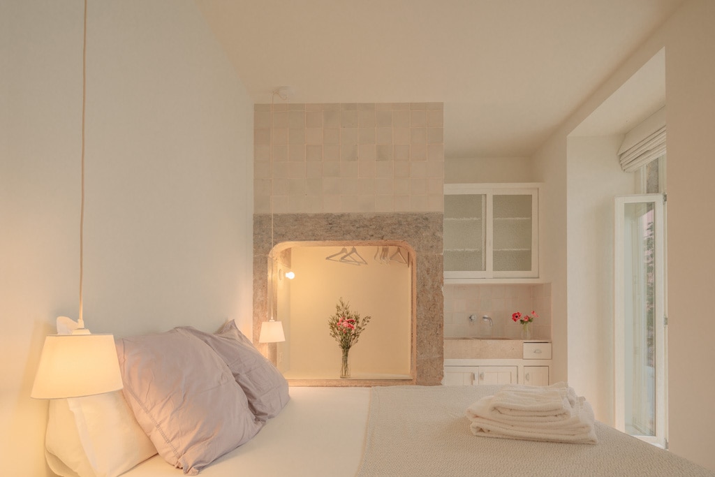 A minimalist bedroom is shown, featuring a spacious bed with soft linens and two bedside lamps. A decorative alcove with a small vase of flowers is visible, along with a light color palette that creates an airy feel. Natural light enters through a nearby window.
