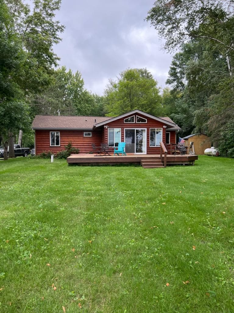 A vibrant red cottage stands amidst lush green grass, framed by trees. The structure features a spacious deck with seating, and large glass doors lead to the interior. The property is set back from the road, providing a private and serene environment.