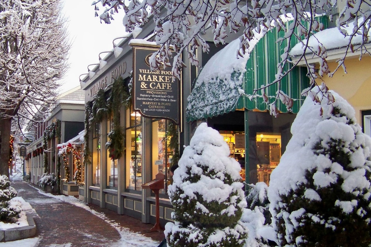 A charming streetscape features a market and café adorned with holiday decorations and snow-covered awnings. Snow-dusted evergreen shrubs are positioned along the pathway, creating a cozy atmosphere in the winter setting, while pedestrians can be seen enjoying the warm ambiance through the large windows.
