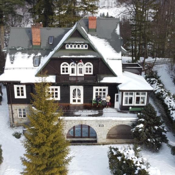 The charming villa is surrounded by snow-covered trees, showcasing a classic design with dark wooden accents. Large windows reflect natural light, and a spacious balcony features a decorative railing. A cozy outdoor area is visible, inviting guests to enjoy the winter landscape.