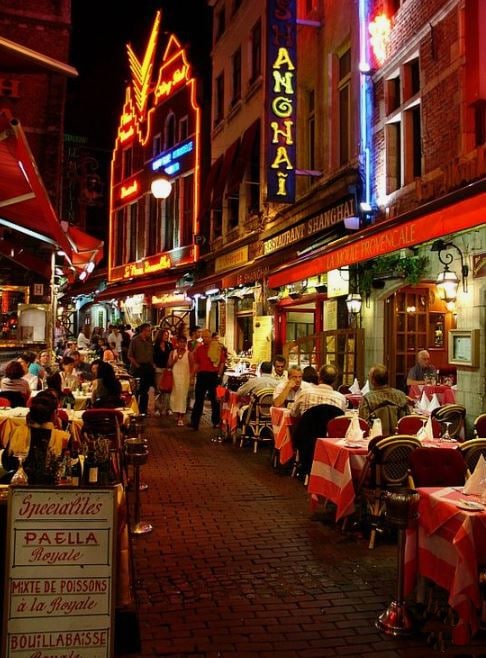 A lively street at night is filled with diners seated at tables adorned with red and white checkered tablecloths. Colorful neon signs illuminate the vibrant atmosphere, while the charming architecture of the historic buildings frames the scene. Patrons are seen enjoying their meals along the bustling walkway.