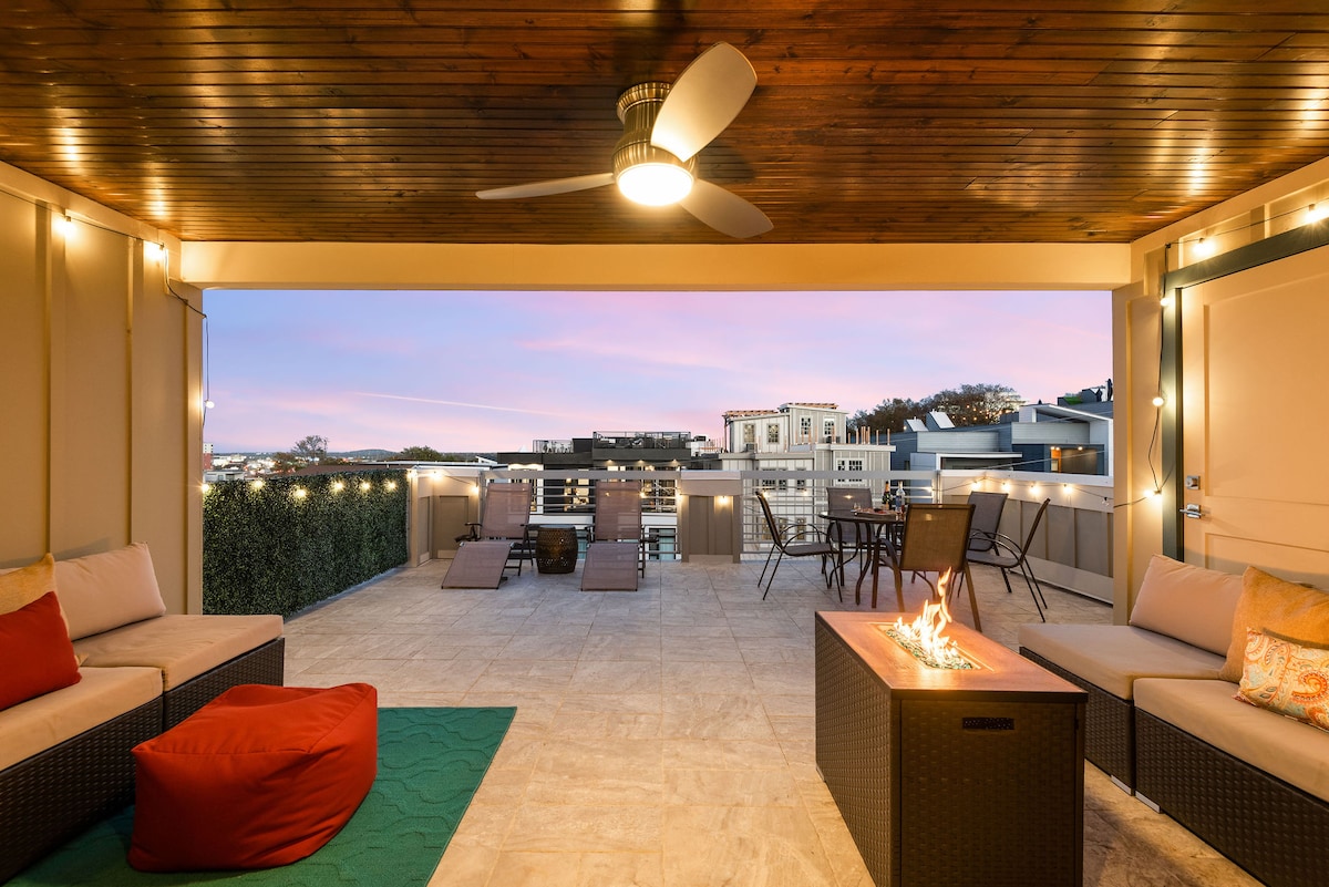An inviting rooftop patio features outdoor sectional seating, a gas fire pit, and sun lounge chairs. Twinkling string lights create a cozy atmosphere, while city skyline views are visible in the background against a colorful sunset.